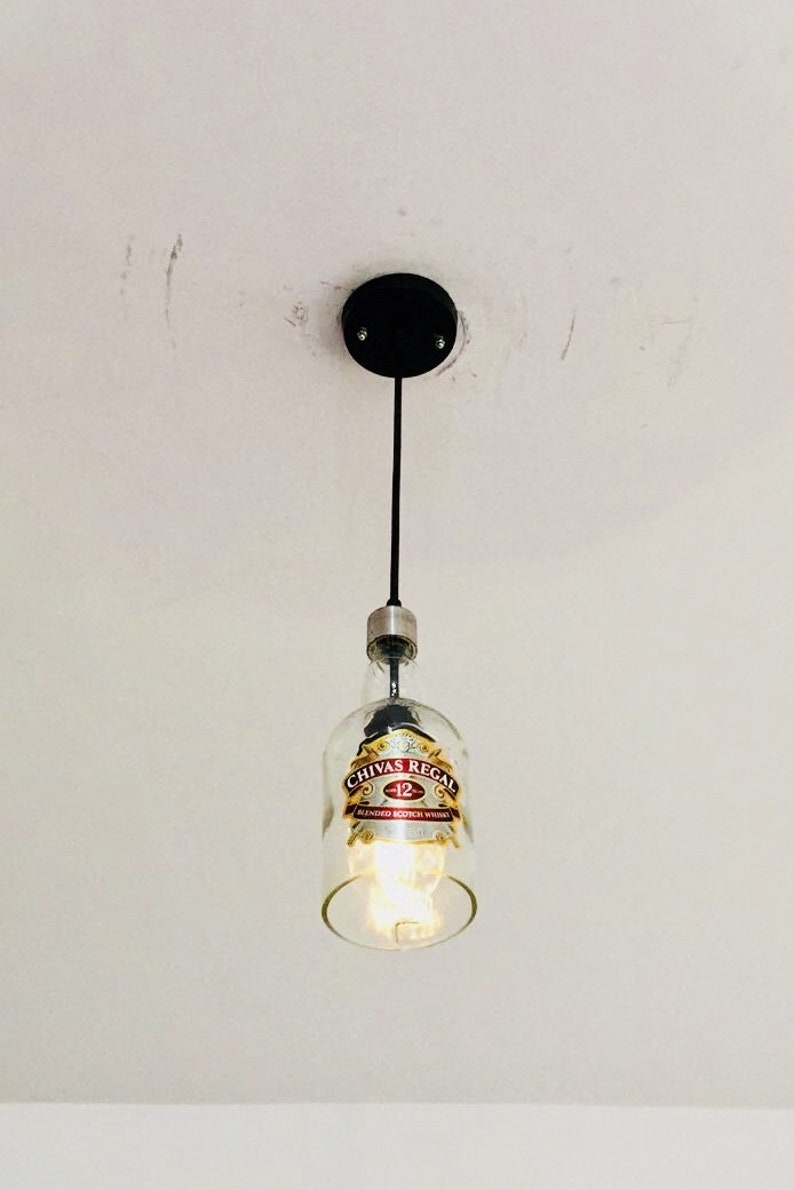 Peut inclure: Un luminaire suspendu r&eacute;alis&eacute; &agrave; partir d'une bouteille de whisky &eacute;cossais m&eacute;lang&eacute; Chivas Regal 12 ans d'&acirc;ge en verre transparent. La bouteille porte une &eacute;tiquette dor&eacute;e avec le logo Chivas Regal et les mots "12" et "Blended Scotch Whisky". Le luminaire est suspendu &agrave; un cordon noir et &agrave; une fixation au plafond noire.