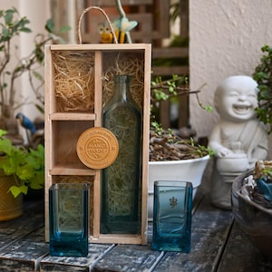 May include: A wooden gift box with a clear glass bottle and two teal-colored square glasses. The box contains decorative straw and a round, gold-colored tag that reads "Hand Made Eco-Friendly Recycled Material". The glasses have a logo on them.
