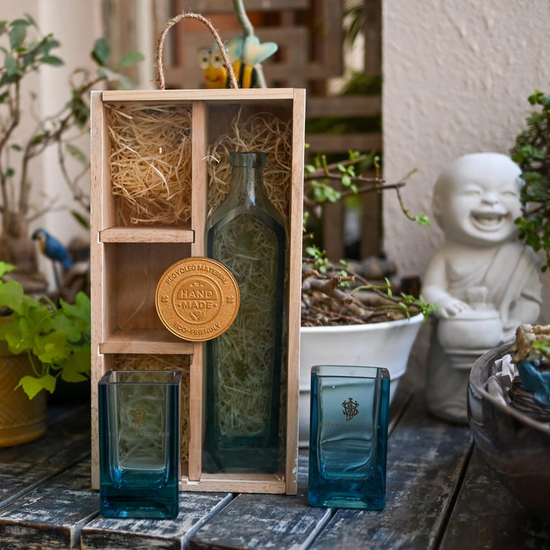 May include: A wooden gift box with a clear glass bottle and two teal-colored square glasses. The box contains decorative straw and a round, gold-colored tag that reads "Hand Made Eco-Friendly Recycled Material". The glasses have a logo on them.