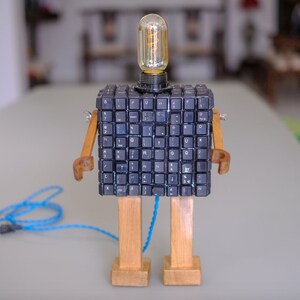 May include: A robot lamp made from a computer keyboard, wooden legs, and a light bulb. The robot is standing on a table with a blue cord.