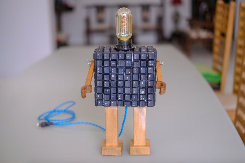 May include: A robot lamp made from a computer keyboard, wooden legs, and a light bulb. The robot is standing on a table with a blue cord.