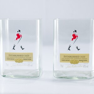 May include: Two clear glass tumblers with a gold label that reads "Established 1820 John Walker & Sons, Kilmarnock, Scotland" and a black and red silhouette of a man walking with a cane. The label also includes the text "Guaranteed  Last Quality" and "Throughout The World".