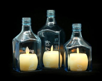 May include: Three clear glass bottles with a light blue tint, each containing a white candle. The bottles have a square shape and are decorated with etched designs.
