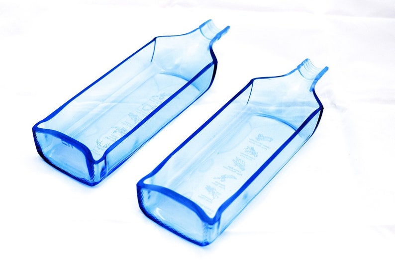 Pu&ograve; includere: Due bottiglie di vetro blu trasparente con un corpo rettangolare e un beccuccio stretto e curvo. Le bottiglie hanno una tinta azzurra e sono poste su uno sfondo bianco. Le bottiglie hanno del testo sul lato.