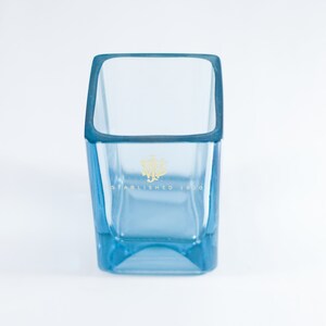 Puede incluir: Un vaso cuadrado de vidrio azul claro con un logotipo dorado que dice "Establecido en 1820".