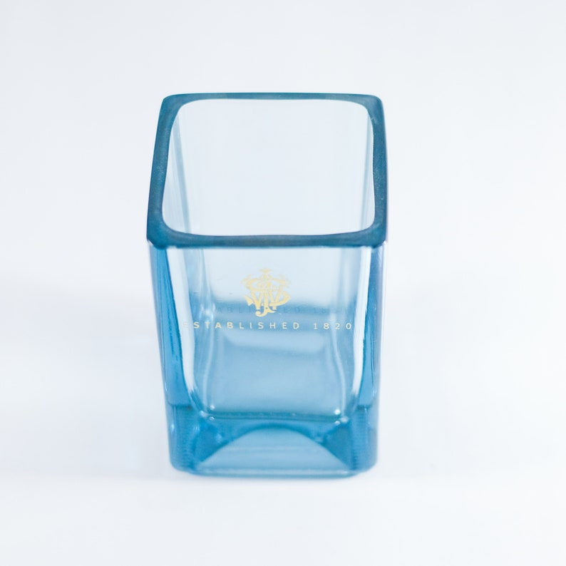 Puede incluir: Un vaso cuadrado de vidrio azul claro con un logotipo dorado que dice "Establecido en 1820".