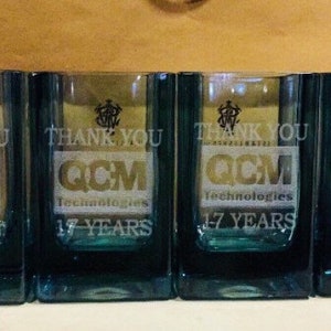 Puede incluir: Cuatro vasos transparentes de vidrio con un dise&ntilde;o esmerilado. Cada vaso tiene el texto "Gracias" y "QCM Technologies 17 a&ntilde;os" grabado en el vidrio.