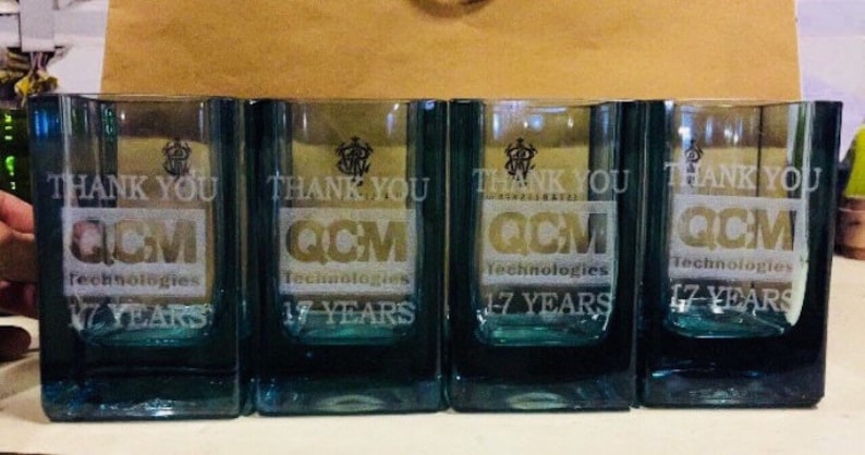 Puede incluir: Cuatro vasos transparentes de vidrio con un dise&ntilde;o esmerilado. Cada vaso tiene el texto "Gracias" y "QCM Technologies 17 a&ntilde;os" grabado en el vidrio.