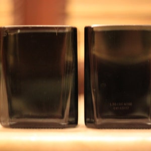 K&ouml;nnte beinhalten: Zwei quadratische Beh&auml;lter aus schwarzem Glas mit glatter Oberfl&auml;che. Der Beh&auml;lter rechts hat ein kleines wei&szlig;es Etikett mit Text darauf.