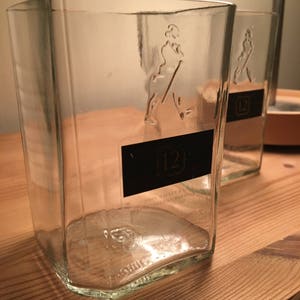 Op de afbeelding: Twee doorzichtige glazen flessen met een getextureerd oppervlak. De flessen zijn leeg en hebben een etiket met de tekst "John Walker & Sons" en "12" erop.