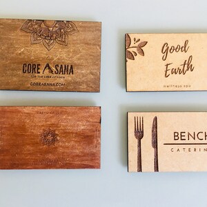 Op de afbeelding: Vier houten visitekaartjes met lasergegraveerde ontwerpen en tekst. De kaart linksboven heeft de tekst "CORE ASANA" met een mandala-ontwerp en de tekst "FOR THE LOVE OF YOGA" en "COREASANA.COM". De kaart rechtsboven heeft de tekst "Good Earth wellness spa" met een bloemmotief. De kaart linksonder heeft een mandala-ontwerp en de tekst "NASHU CHUCH". De kaart rechtsonder heeft de tekst "BENCH CATERING" met een vork- en mesontwerp.