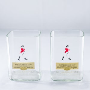 May include: Two clear glass tumblers with a square base. Each glass has a black and white image of a man in a red coat walking with a cane. The text "Established 1820" is printed on each glass.