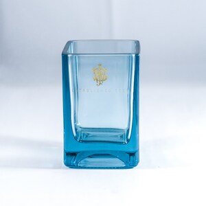 Puede incluir: Un portavelas cuadrado de vidrio transparente con un tono azul. El portavelas tiene un logotipo dorado que dice "Establecido en 1920".