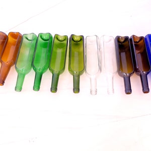 May include: Ten glass bottles, cut in half lengthwise, with the top portion removed. The bottles are arranged in a row, with the cut edges facing the viewer. The bottles are in various colors, including brown, green, clear, and blue.
