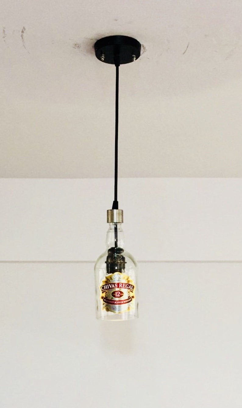 Peut inclure: Un luminaire suspendu en forme de bouteille en verre transparent avec une &eacute;tiquette dor&eacute;e sur laquelle est inscrit "Chivas Regal 12", suspendu &agrave; un cordon noir.