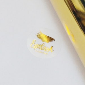 Peut inclure: Un rouleau de papier doré et un autocollant blanc avec des lettres dorées et un motif de plume dorée. L'autocollant indique "Eyelash Extensions" dans une police cursive. Le fond est blanc.
