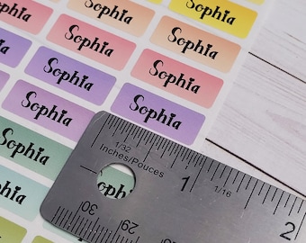 May include: A sheet of colorful rectangular stickers with the name "Sophia" printed in black cursive on each sticker. The stickers are arranged in rows and are approximately 1 inch wide. A ruler is placed on top of the stickers, showing the measurement in inches and 1/32nds of an inch.