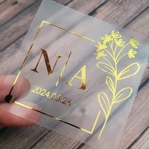 May include: A clear, square sticker with a gold border and gold lettering. The sticker features the initials "N" and "A" with a floral design and the date "2024.05.26".