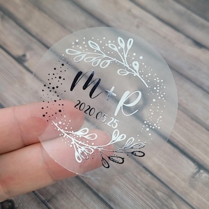 May include: Clear round sticker with a silver floral design and black lettering. The sticker says "M+R 2020.05.25".