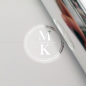 May include: A silver sticker with a circular design featuring the letters "M" and "K" in a monogram style.