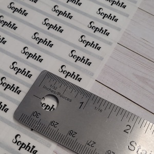 May include: A sheet of white labels with the name "Sophia" printed in black cursive on each label. A ruler is placed on top of the labels, showing measurements in inches.