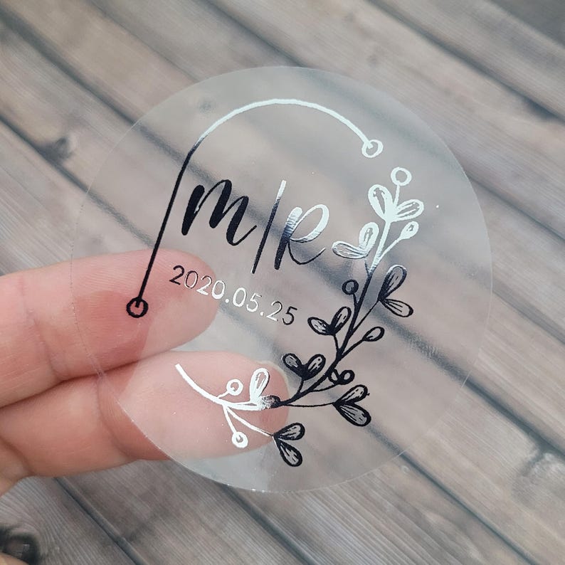 Puede incluir: Pegatina redonda transparente con fondo plateado y letras negras. La pegatina dice "m|k" y "2020.05.25" con un dise&ntilde;o floral.