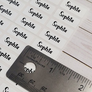 May include: A close-up of a ruler measuring white rectangular stickers with the name "Sophia" printed in black cursive lettering.
