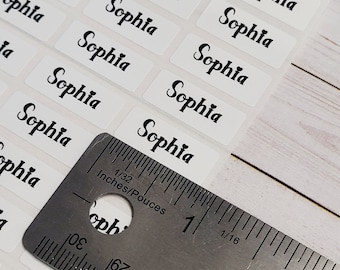 May include: A close-up of a ruler measuring white rectangular stickers with the name "Sophia" printed in black cursive lettering.