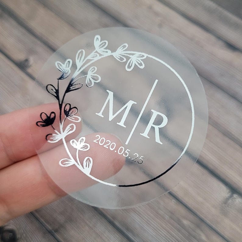 Puede incluir: Pegatina redonda transparente con un dise&ntilde;o de corona floral plateada y las letras "M/R" y la fecha "2020.05.25" en plata.
