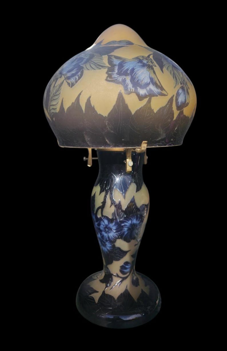 May include: An antique table lamp with a floral design. The lamp features a beige and blue glass shade with a dark blue base. The shade and base are decorated with blue flowers and dark blue leaves. The lamp is a decorative lighting fixture.