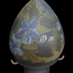 May include: A decorative glass lamp with a floral design. The lamp features a teardrop-shaped shade with blue flowers and green leaves against a yellow background. The base is a matching color and design. The lamp has two curved handles.