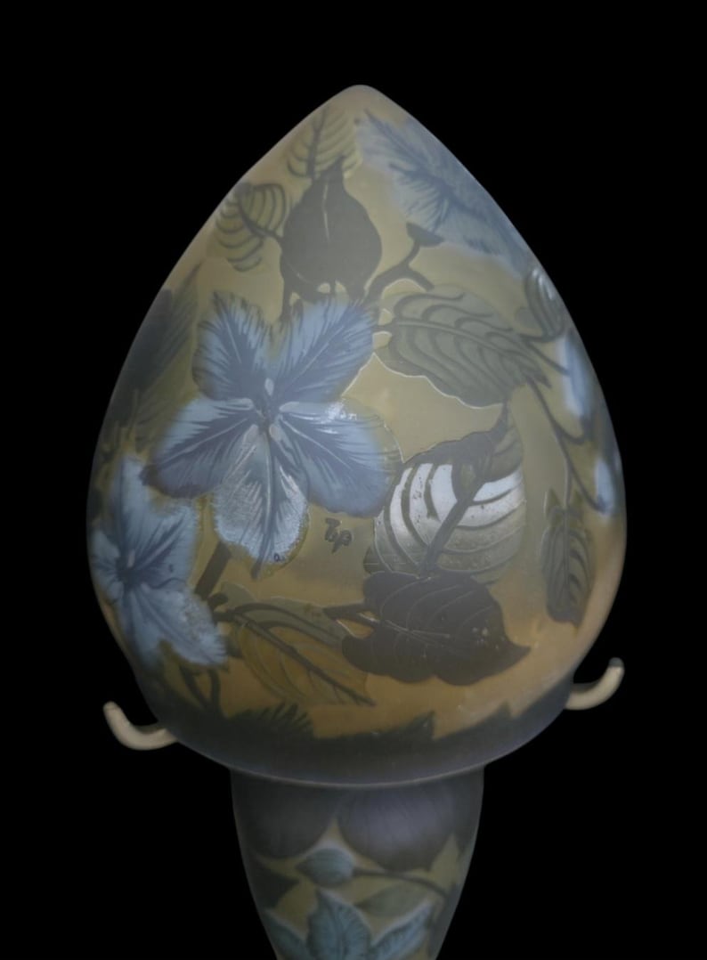 May include: A decorative glass lamp with a floral design. The lamp features a teardrop-shaped shade with blue flowers and green leaves against a yellow background. The base is a matching color and design. The lamp has two curved handles.