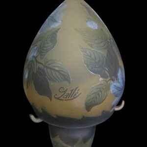 May include: An art glass lamp with a teardrop-shaped shade. The lamp features a botanical design of leaves in shades of green and blue against a yellow background. The lamp has the signature "Gall&eacute;" etched on the front.