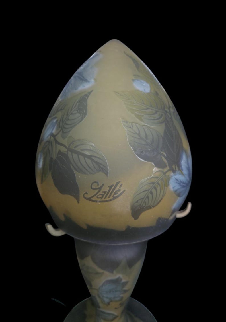 May include: An art glass lamp with a teardrop-shaped shade. The lamp features a botanical design of leaves in shades of green and blue against a yellow background. The lamp has the signature "Gall&eacute;" etched on the front.
