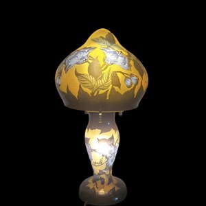 May include: A decorative table lamp with a yellow, mushroom-shaped shade featuring floral and leaf designs in shades of gray and brown. The lamp's base is a matching color and design, with a curved stem and a round base. The lamp is lit from within.