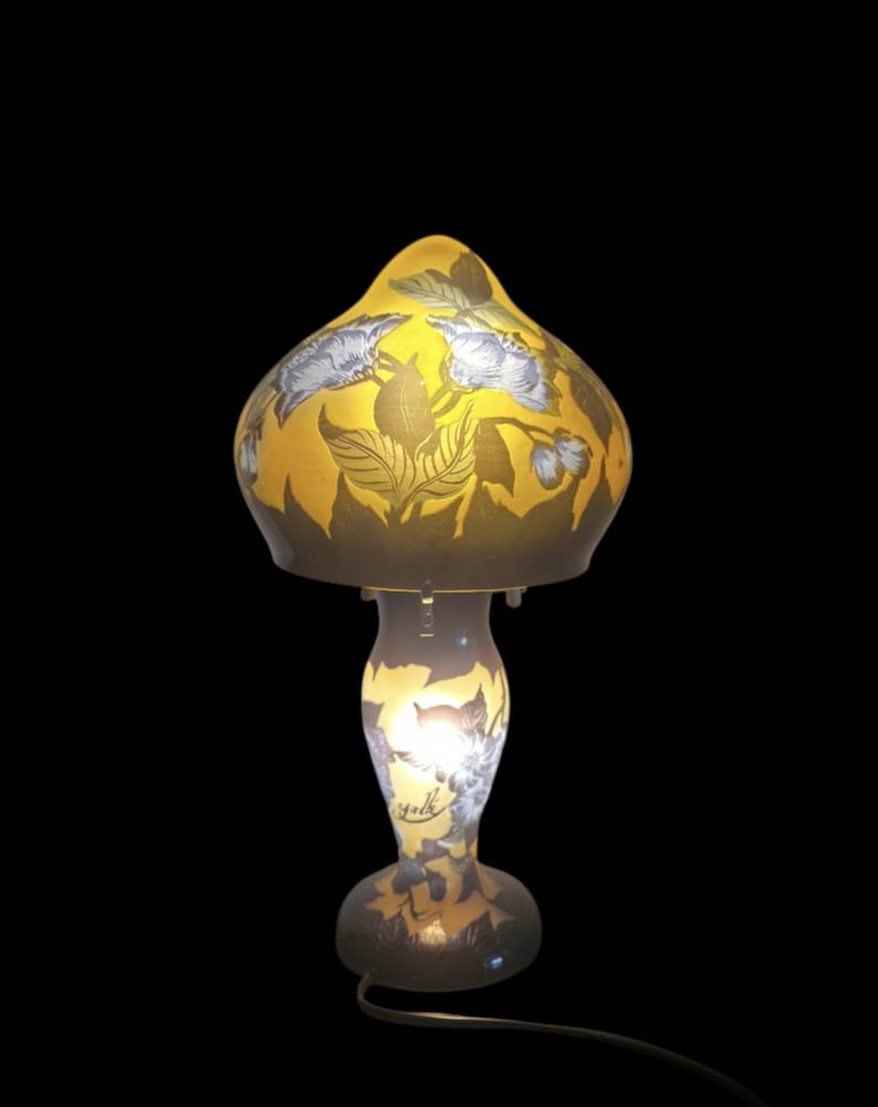 May include: A decorative table lamp with a yellow, mushroom-shaped shade featuring floral and leaf designs in shades of gray and brown. The lamp's base is a matching color and design, with a curved stem and a round base. The lamp is lit from within.