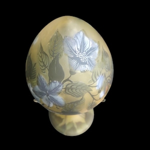 May include: An egg-shaped glass lamp with a floral pattern. The lamp has a pale yellow colour with blue and white flowers and green leaves. It stands on a round base. The background is black.