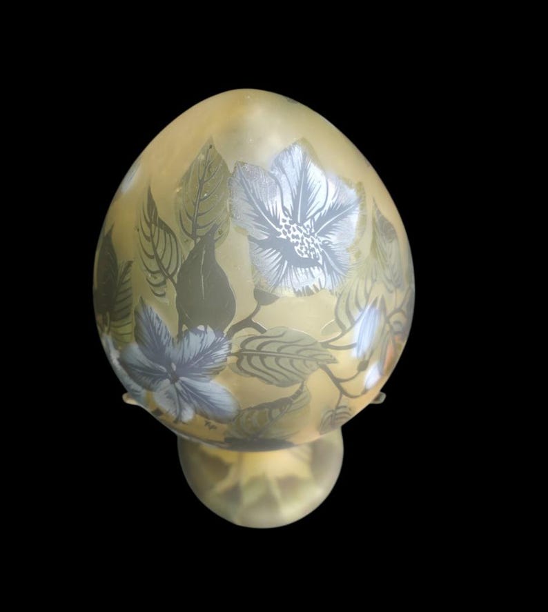May include: An egg-shaped glass lamp with a floral pattern. The lamp has a pale yellow colour with blue and white flowers and green leaves. It stands on a round base. The background is black.