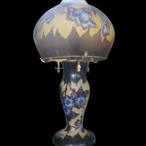 May include: An antique table lamp with a floral design. The lamp features a dark blue base and shade with a gold-colored background. The shade has a rounded shape and is decorated with blue flowers and leaves. The lamp is made of glass and metal.