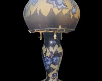 May include: An antique table lamp with a floral design. The lamp has a dark blue base and shade with a gold-coloured background. The shade has a rounded shape and is decorated with blue flowers and leaves. The lamp is made of glass and metal.