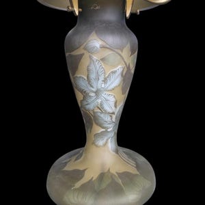 May include: An antique art nouveau lamp with a floral design. The glass base is a light tan colour with etched leaves and a large blue-grey flower. The lamp has a brass-coloured metal frame and a dark shade.