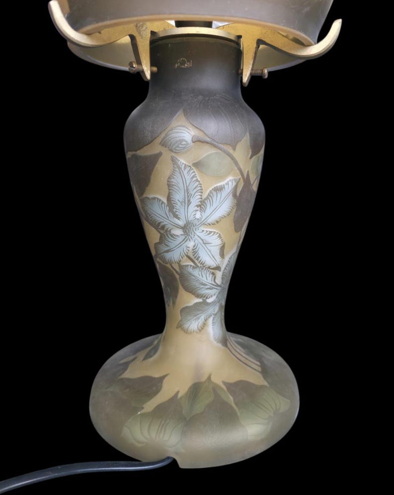 May include: An antique art nouveau lamp with a floral design. The glass base is a light tan colour with etched leaves and a large blue-grey flower. The lamp has a brass-coloured metal frame and a dark shade.