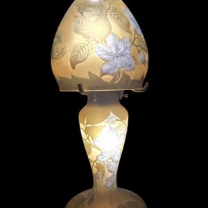 May include: A decorative table lamp with a floral design. The lamp features a light yellow glass shade and base, adorned with white and grey floral and leaf patterns. The lamp has a curved, elongated shape and is set against a black background.