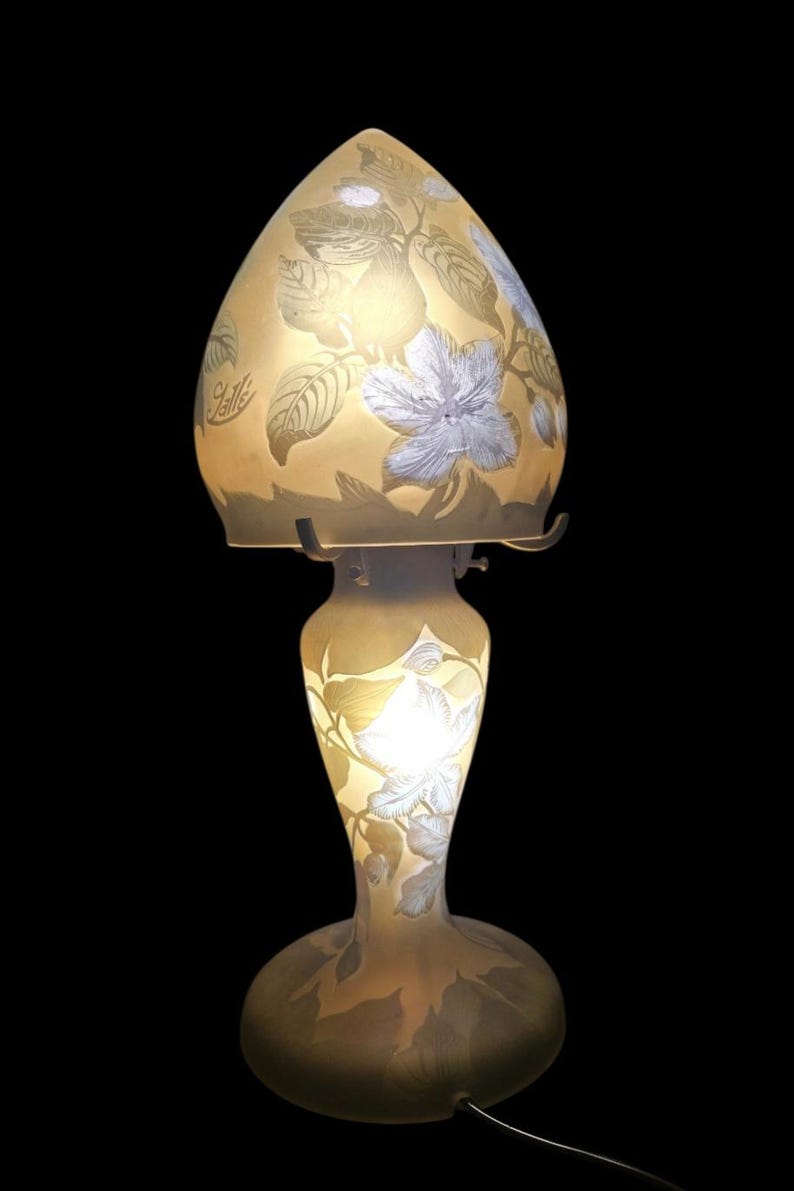 May include: A decorative table lamp with a floral design. The lamp features a light yellow glass shade and base, adorned with white and grey floral and leaf patterns. The lamp has a curved, elongated shape and is set against a black background.