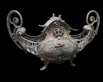 May include: An ornate silver-coloured centerpiece bowl with intricate scrollwork and floral designs. The bowl has a boat-like shape with curved handles and a decorative lattice pattern. It rests on three small feet.