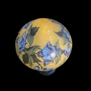 May include: A round, yellow glass decorative object with blue floral designs and green leaves. The object has a dark blue base and is set against a black background. The floral pattern is detailed and covers the entire surface.