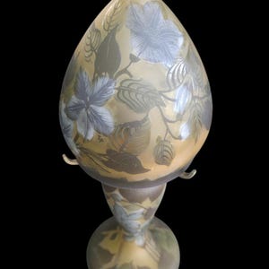 May include: A decorative table lamp with a teardrop-shaped shade. The lamp features a floral design in shades of blue and green on a yellow background. The base is round and the lamp has a black cord.