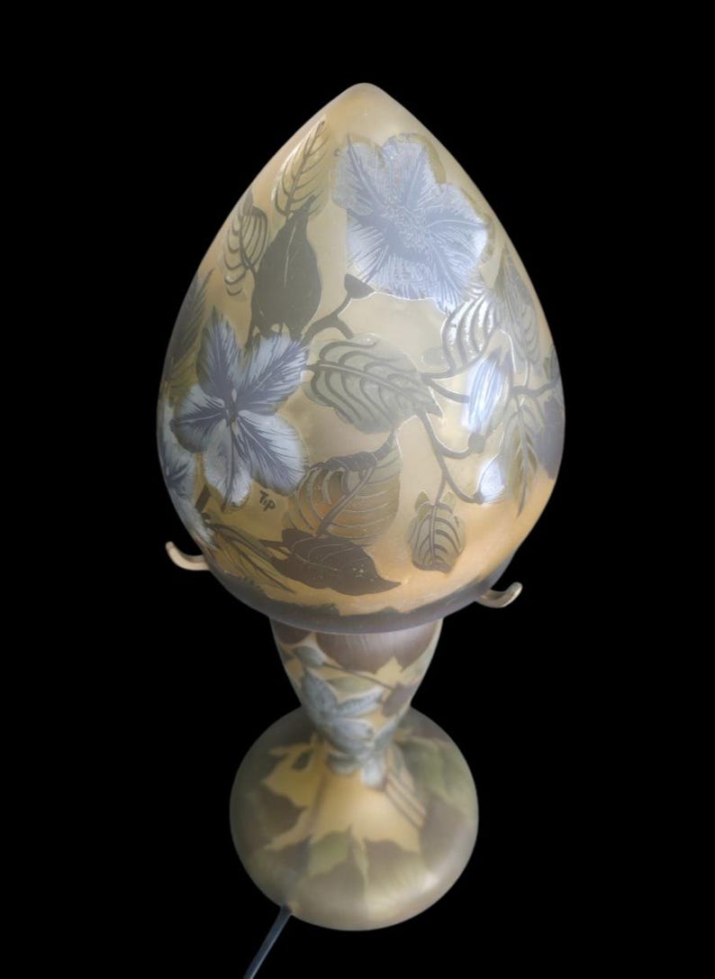 May include: A decorative table lamp with a teardrop-shaped shade. The lamp features a floral design in shades of blue and green on a yellow background. The base is round and the lamp has a black cord.