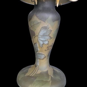 May include: An antique art nouveau lamp with a frosted glass shade and a decorative base. The base features a floral design with the word "Gall&eacute;" in script. The lamp has a brass-coloured metal frame and a dark background.