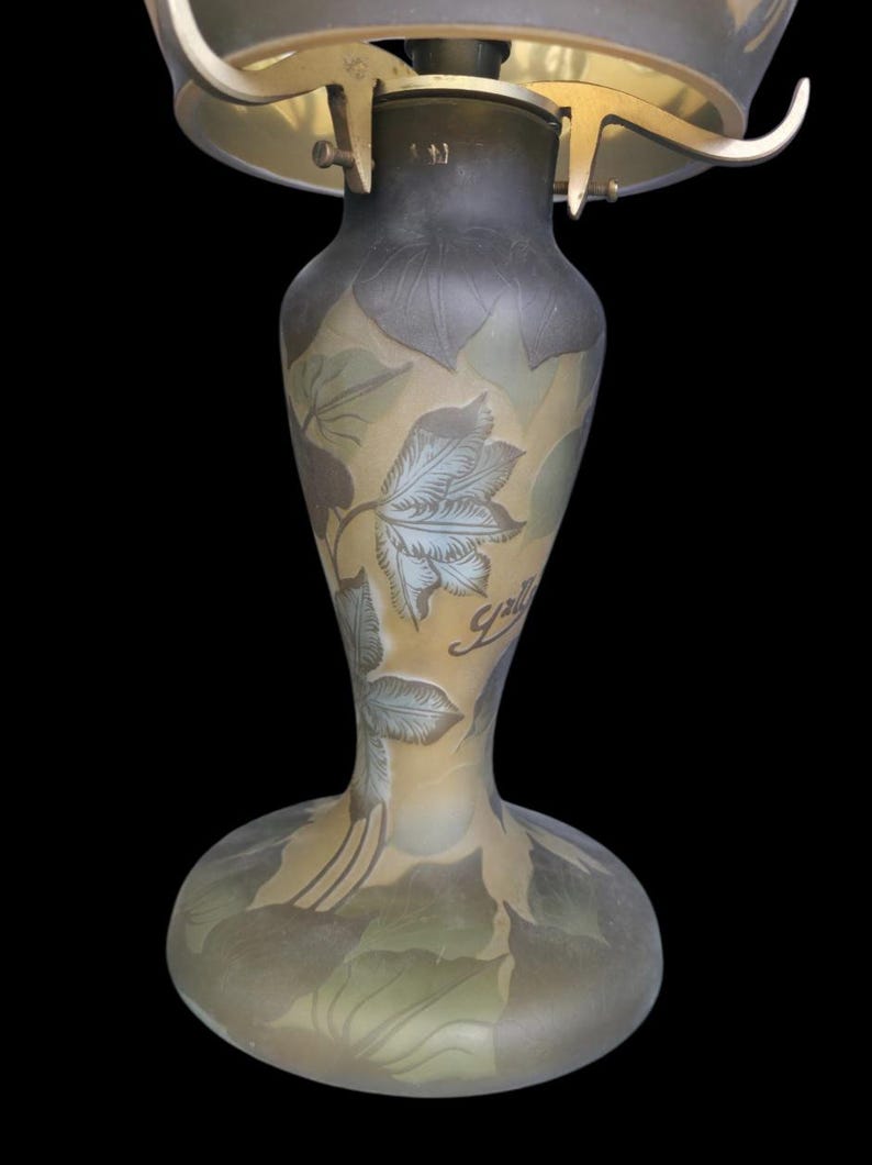 May include: An antique art nouveau lamp with a frosted glass shade and a decorative base. The base features a floral design with the word "Gall&eacute;" in script. The lamp has a brass-coloured metal frame and a dark background.